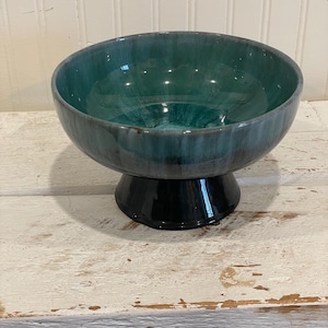 May include: A teal and black ceramic bowl with a pedestal base. The bowl has a glossy finish and a wide, open design. The interior of the bowl is a lighter shade of teal, creating a gradient effect. The base is black.
