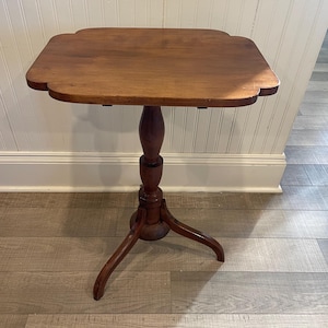 May include: A small, antique wooden side table with a scalloped edge tabletop. The table is supported by a turned pedestal and three curved legs. The wood has a warm, reddish-brown tone.