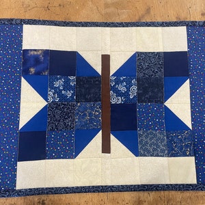May include: A blue and white patchwork quilt with a butterfly design in the centre. The quilt is made of various blue fabrics with different patterns, including floral, paisley, and geometric designs. The butterfly is made of a brown fabric and is surrounded by a white border.