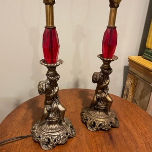 Puede incluir: Par de lámparas de mesa antiguas con accesorios de latón, detalles de cristal rojo facetado y base de querubín ornamentada. Las lámparas tienen una estética vintage y son adecuadas para una sala de estar o dormitorio. Las lámparas miden aproximadamente 51 cm de alto.