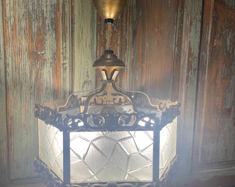 Puede incluir: Una lámpara de techo vintage de latón y vidrio con una pantalla de vidrio con patrón geométrico. La lámpara tiene un diseño decorativo y ornamentado y está colgada de una cadena.