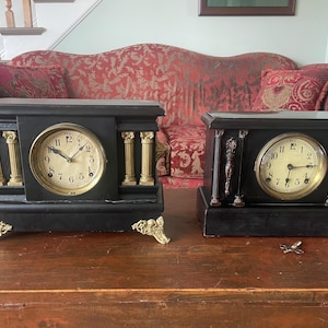 May include: Two antique black mantel clocks with gold accents. The clocks have round faces with Roman numerals and ornate gold detailing. One clock has gold columns, and both sit on a wooden surface.