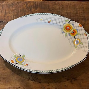 May include: An oval white ceramic serving platter featuring a decorative border with a thin green striped edge. The surface is adorned with a black grid pattern, small orange and yellow squares, and stylised floral motifs in yellow, orange, and blue.