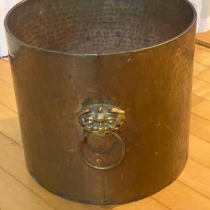 Könnte beinhalten: Zylindrischer Kupferbehälter mit gehämmertem Muster. Ein dekorativer Löwenkopf mit Ringgriff ist an der Seite befestigt. Der Eimer hat eine warme, metallisch braune Farbe und steht auf einem Holzboden.
