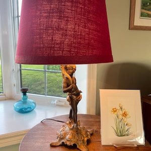 Peut inclure: Une lampe de table décorative avec un abat-jour bordeaux. Le pied de la lampe représente un chérubin de couleur bronze. Une aquarelle encadrée de jonquilles et une petite boîte à bijoux ronde à couvercle sont sur la table.