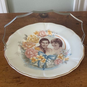 May include: A white ceramic plate with a gold rim features a floral wreath surrounding a photo of Prince Charles and Lady Diana Spencer. The plate has a silver handle.
