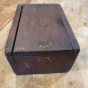May include: A rectangular wooden box with a dark brown finish and a hinged lid. The surface of the wood shows signs of wear, including scratches and scuffs, with a small rectangular patch of darker material visible on the top of the lid.