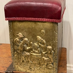 May include: A brass coal scuttle with a red leather top and an embossed design of people gathered around a barrel. The scuttle has a decorative border around the top.