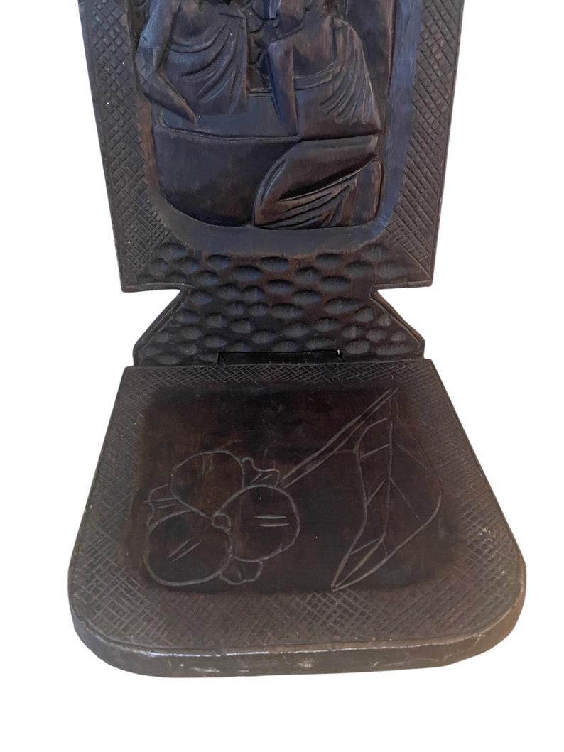 May include: A dark brown wooden folding chair with intricate carvings of figures and a floral design on the seat.