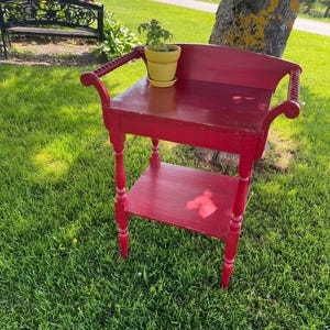 May include: A distressed red wooden side table with a lower shelf and curved handles. A yellow plant pot sits on the top. The table is on a grassy lawn. The table's height is approximately 76 cm.