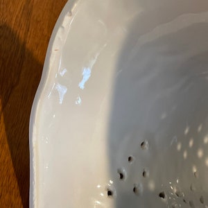 May include: White ceramic bowl with a scalloped edge and small holes in the bottom. The bowl is designed for straining liquids.