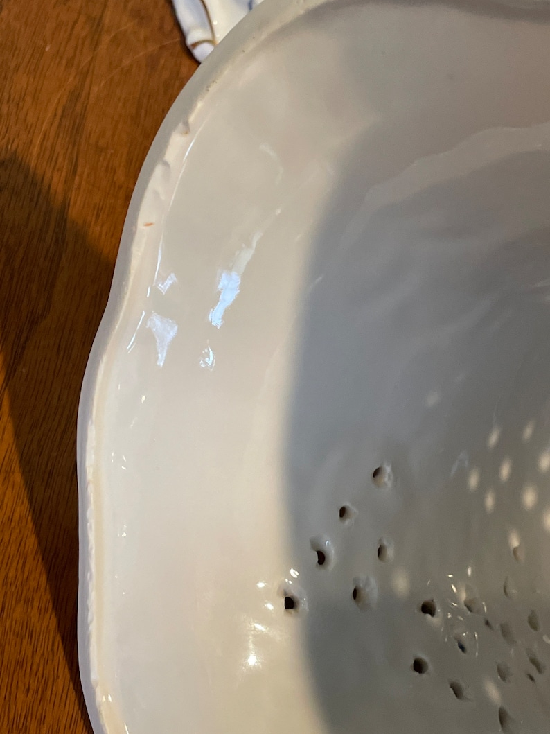 May include: White ceramic bowl with a scalloped edge and small holes in the bottom. The bowl is designed for straining liquids.