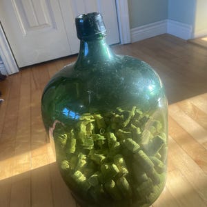 Puede incluir: Una botella grande de vidrio verde llena de objetos cilíndricos verdes. La botella tiene una tapa negra y está parcialmente llena de un líquido verde claro. Los objetos del interior parecen estar muy juntos. La botella está sobre una superficie de madera.