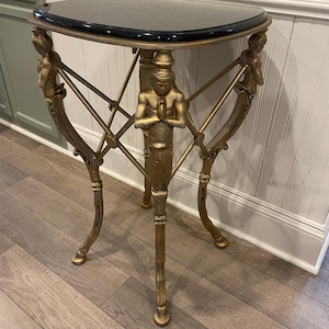 May include: A gold-coloured, three-legged accent table with a black, semi-circular top. The table features ornate, stylized figures and geometric supports. The table is approximately 76 cm tall.