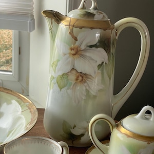 Puede incluir: Un juego de té de porcelana vintage con un diseño floral. El juego incluye una tetera, una jarra de crema, un azucarero y una taza de té. El diseño floral presenta flores blancas con hojas verdes sobre un fondo crema. El juego está adornado con detalles dorados.