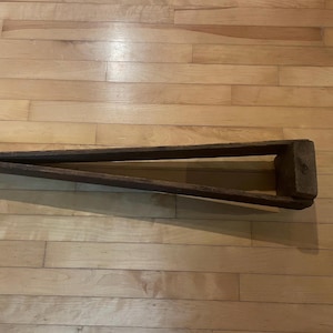 May include: A wooden tool with a triangular shape and two arms. The tool is made of dark brown wood and has a rough texture.