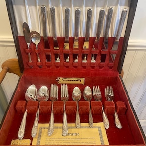 May include: A vintage silverware set in a wooden case with a red velvet interior. The set includes spoons, forks, and knives with ornate handles. The case has a Community label and a guarantee card.