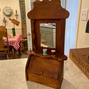 May include: A vintage wooden sewing cabinet with a mirror, featuring a small drawer and a shelf holding two spools of thread. The cabinet has a dark brown finish and a decorative top, perfect for storing sewing supplies.