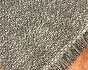 May include: A gray woven table runner with a fringe border. The runner has a subtle herringbone pattern.