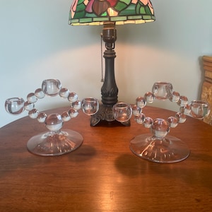 May include: Two clear glass candle holders with a circular design of small glass spheres and candle cups. The candle holders sit on a round base. A lamp with a stained glass shade is in the background.