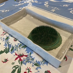 May include: A white ceramic rectangular tray with a green floral foam insert. The tray is on a white and blue floral tablecloth.