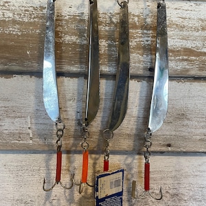 May include: Four vintage fishing lures hanging on a wooden wall. The lures are made of shiny metal with a curved shape, each connected to a treble hook with a red or orange plastic tube. A blue and white tag hangs from one lure.