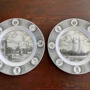May include: Two decorative plates with black and white nautical scenes. Each plate features a central image of a harbor or ship, surrounded by a gray border with nautical symbols. The plates are on a dark wooden surface.