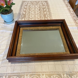 Puede incluir: Un espejo rectangular con un marco de madera de color marrón oscuro y un borde interior texturizado de color dorado. El espejo tiene una superficie de vidrio clara y reflectante, encajada en el marco multicapa con acabado de madera.