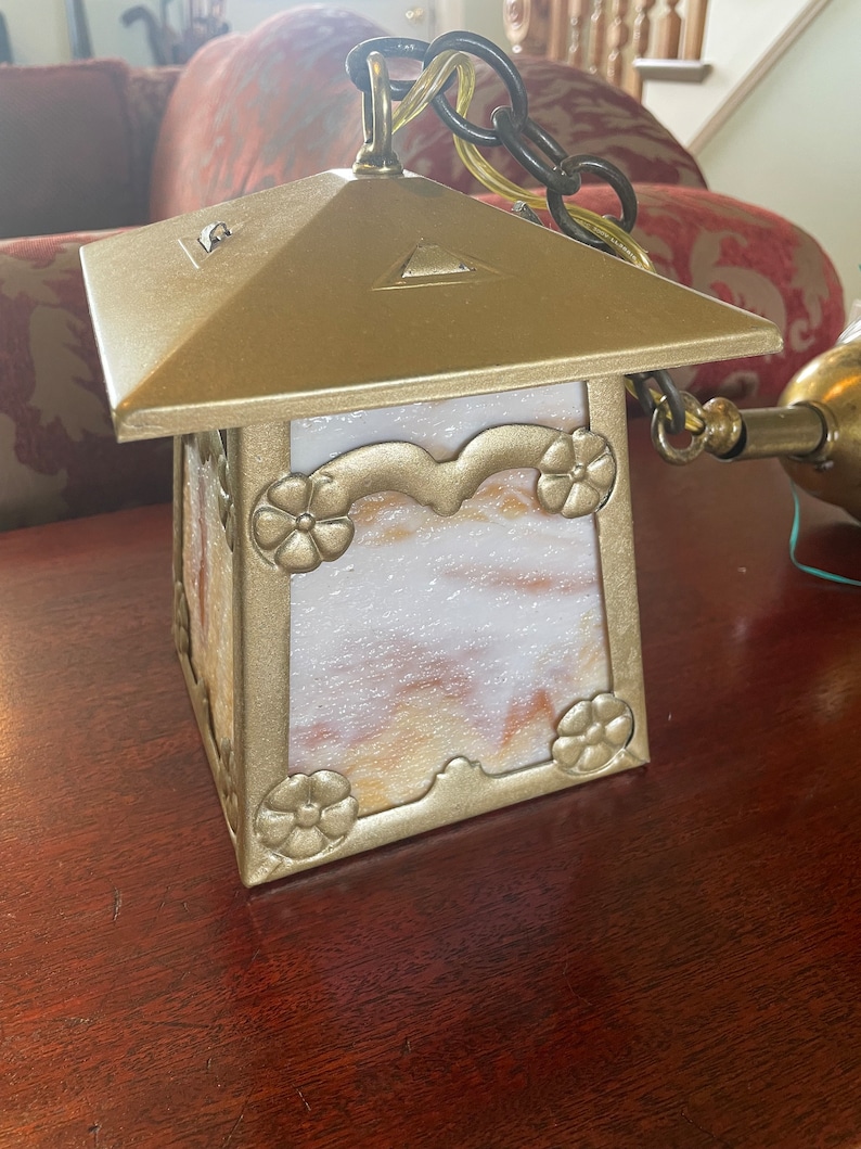May include: A gold-toned metal hanging light fixture with a square, frosted glass shade featuring a floral design. The shade has a white and brown marbled pattern.