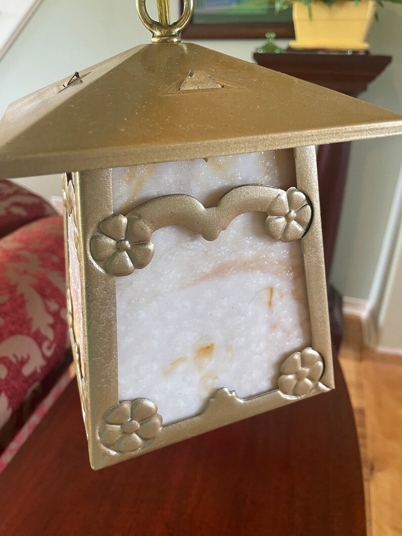 May include: A gold-toned pendant light fixture with a square, frosted glass shade. The shade has a decorative floral design around the edges and a white, marbled pattern.