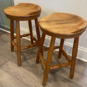 May include: Two wooden bar stools with a medium brown finish. Each stool features a round, contoured seat with a visible wood grain pattern and four legs connected by decorative, turned-style stretchers that provide structural support.