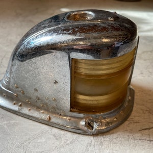 May include: A vintage chrome and glass boat navigation light. The light has a rounded, dome-shaped top and a rectangular lens with multiple horizontal ridges. The lens is clear and has a yellow tint.