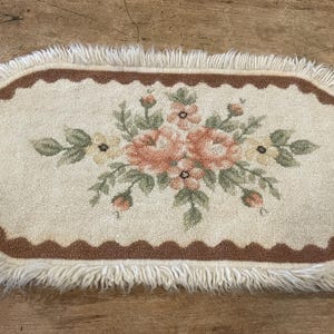 May include: An oval-shaped rug with a cream-colored center featuring a floral design in shades of peach, pink, and yellow. The rug has a brown scalloped border and a fringe edge. The rug is likely made of a soft, woven material.