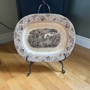Könnte beinhalten: Eine große, ovale, cremefarbene Keramik-Servierplatte mit einer braunen und weißen Landschaftsszene. Die Platte steht auf einem schwarzen Metallständer mit drei Beinen.
