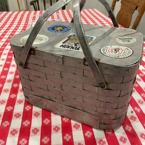 May include: A silver metal picnic basket with a woven design and a handle. The basket has several vintage stickers on the top.