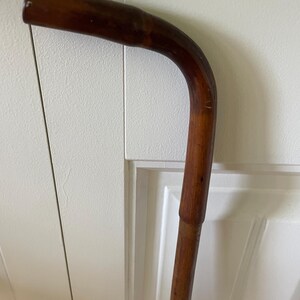 May include: A dark brown wooden walking cane featuring a curved handle. The wood displays a natural grain pattern with a smooth, polished finish. The cane has a cylindrical shaft that connects to a rounded, ergonomic grip at the top end.