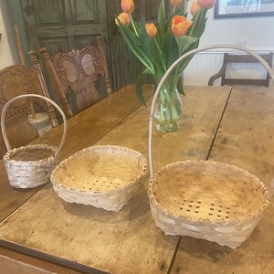 May include: Three woven baskets of different sizes on a wooden table. The baskets are light-coloured and have handles. A vase of orange tulips is in the background. The table and chairs have a rustic, antique appearance.