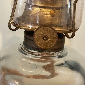 May include: Close-up of a vintage oil lamp with a glass chimney and a brass burner. The lamp has a round, clear glass reservoir and a metal base with a label that reads "VERA CO. PAT. APPL'D FOR".