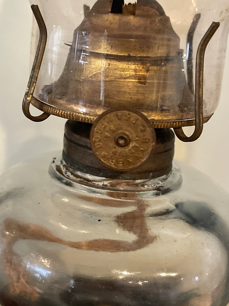 May include: Close-up of a vintage oil lamp with a glass chimney and a brass burner. The lamp has a round, clear glass reservoir and a metal base with a label that reads "VERA CO. PAT. APPL'D FOR".