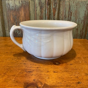 May include: White ceramic chamber pot with a single handle. The pot has a fluted design and a raised floral pattern.