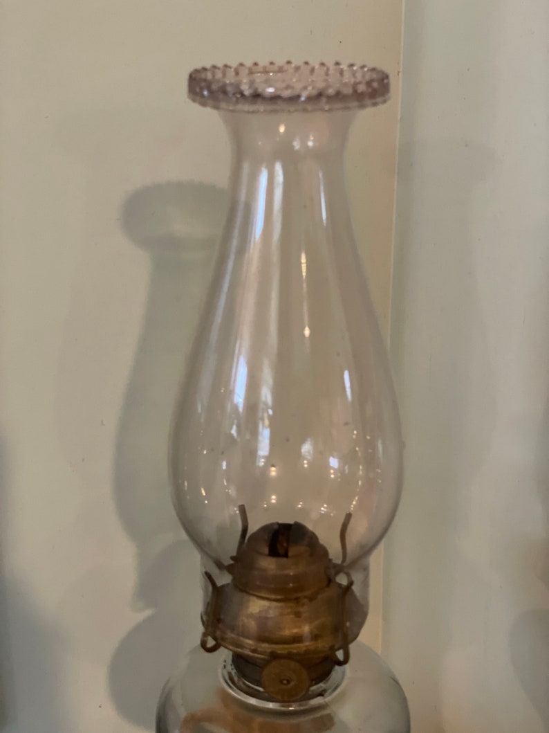 May include: A clear glass oil lamp with a frosted glass chimney and a metal burner. The chimney has a beaded rim.