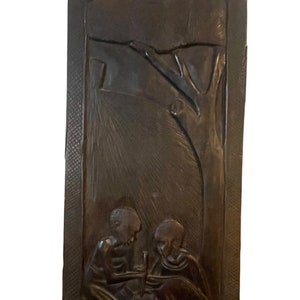 May include: A dark brown wooden wall hanging with a carved scene of two people sitting under a tree. The carving is detailed and shows the people's faces and clothing.