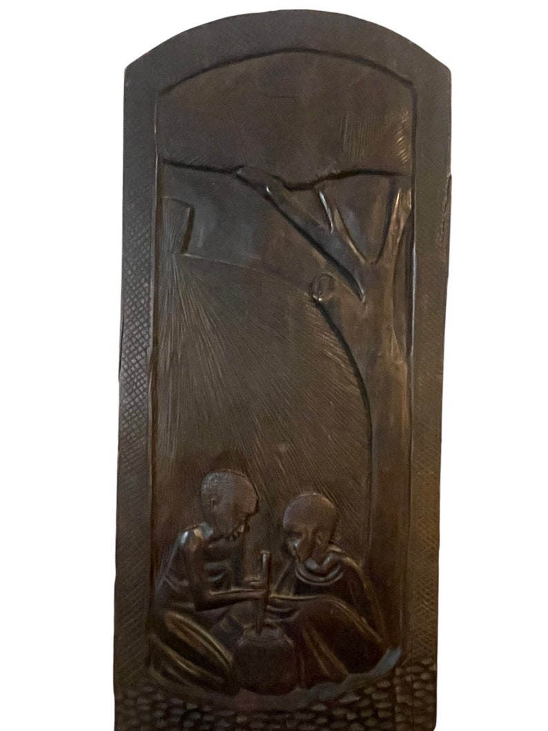 May include: A dark brown wooden wall hanging with a carved scene of two people sitting under a tree. The carving is detailed and shows the people's faces and clothing.
