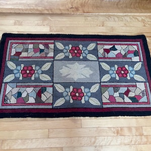 May include: A vintage rug with a geometric design featuring red, blue, and green flowers and a stained glass pattern. The rug has a black border and a burgundy and cream background.