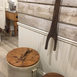 May include: Two white ceramic crocks with wooden lids. The crocks are sitting on a wooden floor. A wooden utensil hangs on a white wall behind the crocks.