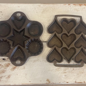 Peut inclure: Deux moules à pâtisserie anciens en fonte. L'un présente diverses formes, dont un cœur, une étoile et des cercles. L'autre est en forme de sapin de Noël avec des cavités en forme de cœur. Les deux moules sont brun foncé et reposent sur une surface en bois blanc.