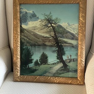May include: A rectangular landscape painting in a decorative gold-toned frame with floral relief details. The artwork depicts a serene mountain lake with snow-capped peaks, a prominent leaning tree in the foreground, and two small figures walking.