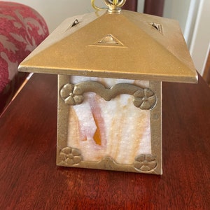 May include: A brass lantern with a square base and a square top. The lantern has a clear glass panel with a floral design and a white and orange marbled pattern. The lantern has a chain hanging from the top.