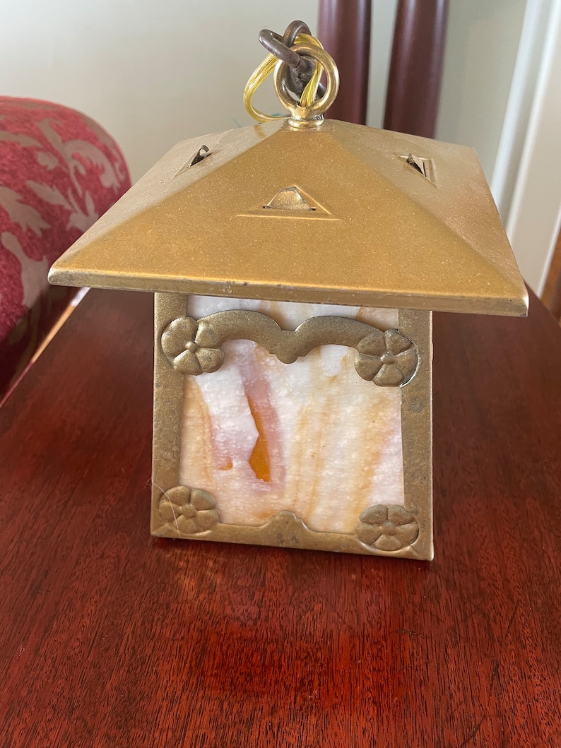 May include: A brass lantern with a square base and a square top. The lantern has a clear glass panel with a floral design and a white and orange marbled pattern. The lantern has a chain hanging from the top.