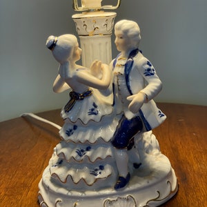 May include: A white porcelain table lamp with a blue and white floral design. The lamp features a couple dancing, with the woman in a white dress and the man in a blue and white suit.